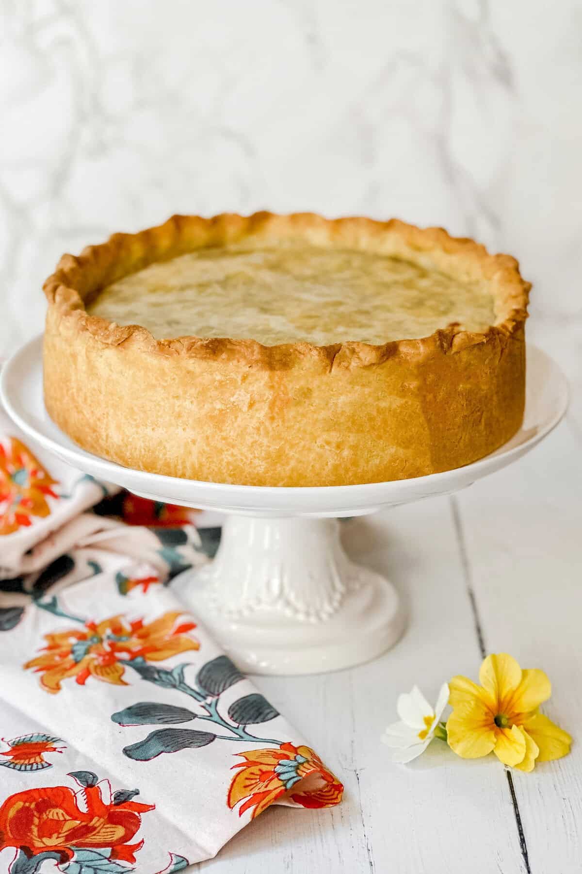 A deep-dish pie, reminiscent of a classic pizza rustica, boasts a golden-brown crust on a white cake stand. A floral-patterned cloth and yellow and white flowers decorate the white wooden surface, set against a light marble-textured background.