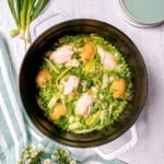 A white pot filled with Spring Chicken, potatoes, peas, green onions, and herbs sits on a white wooden surface with plates and flowers nearby.