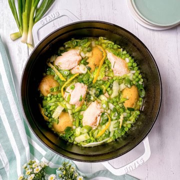 A white pot filled with Spring Chicken, potatoes, peas, green onions, and herbs sits on a white wooden surface with plates and flowers nearby.