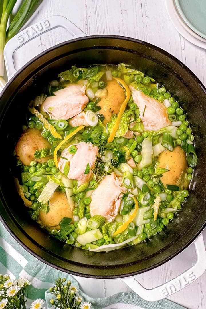 A white pot filled with Spring Chicken, potatoes, peas, green onions, and herbs sits on a white wooden surface with plates and flowers nearby.