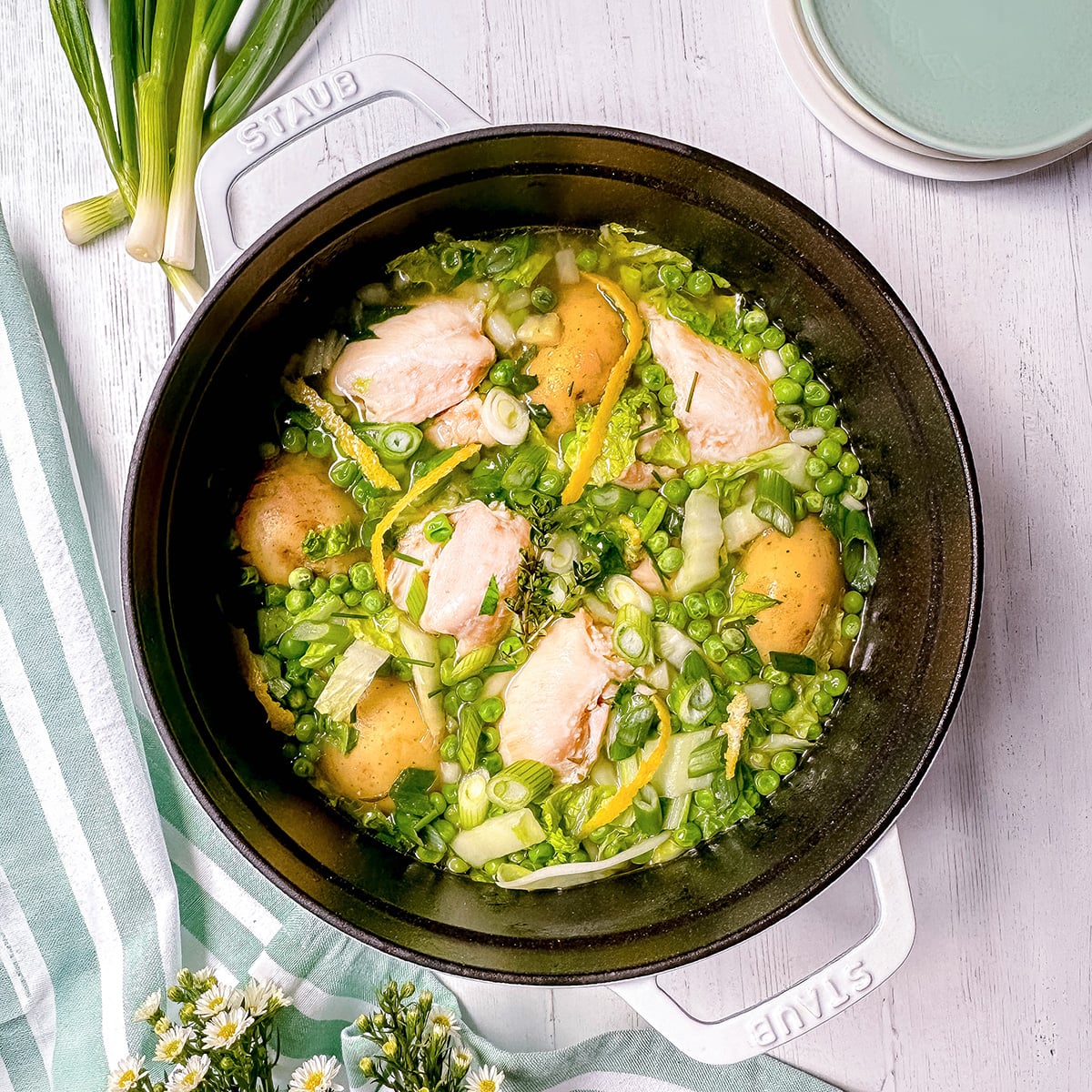 A white pot filled with Spring Chicken, potatoes, peas, green onions, and herbs sits on a white wooden surface with plates and flowers nearby.