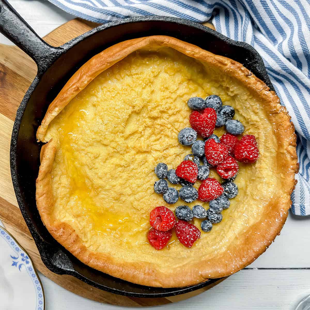 Square image of baked German Pancake in a cast iron skillet topped with fresh blueberries and raspberries.