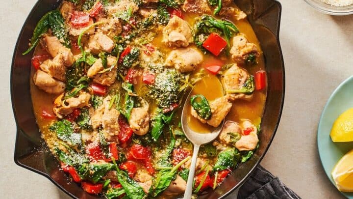 A skillet filled with cooked chicken, spinach, and red bell peppers in a light sauce, perfect for March 400 Calorie Dinners, with a spoon beside a bowl of grated cheese and lemon wedges.