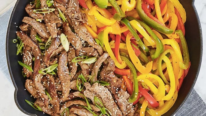 A skillet with cooked beef strips on one side and sliced red, yellow, and green bell peppers on the other, garnished with sesame seeds and green onions—perfect for March Healthy Comfort Food Dinners.