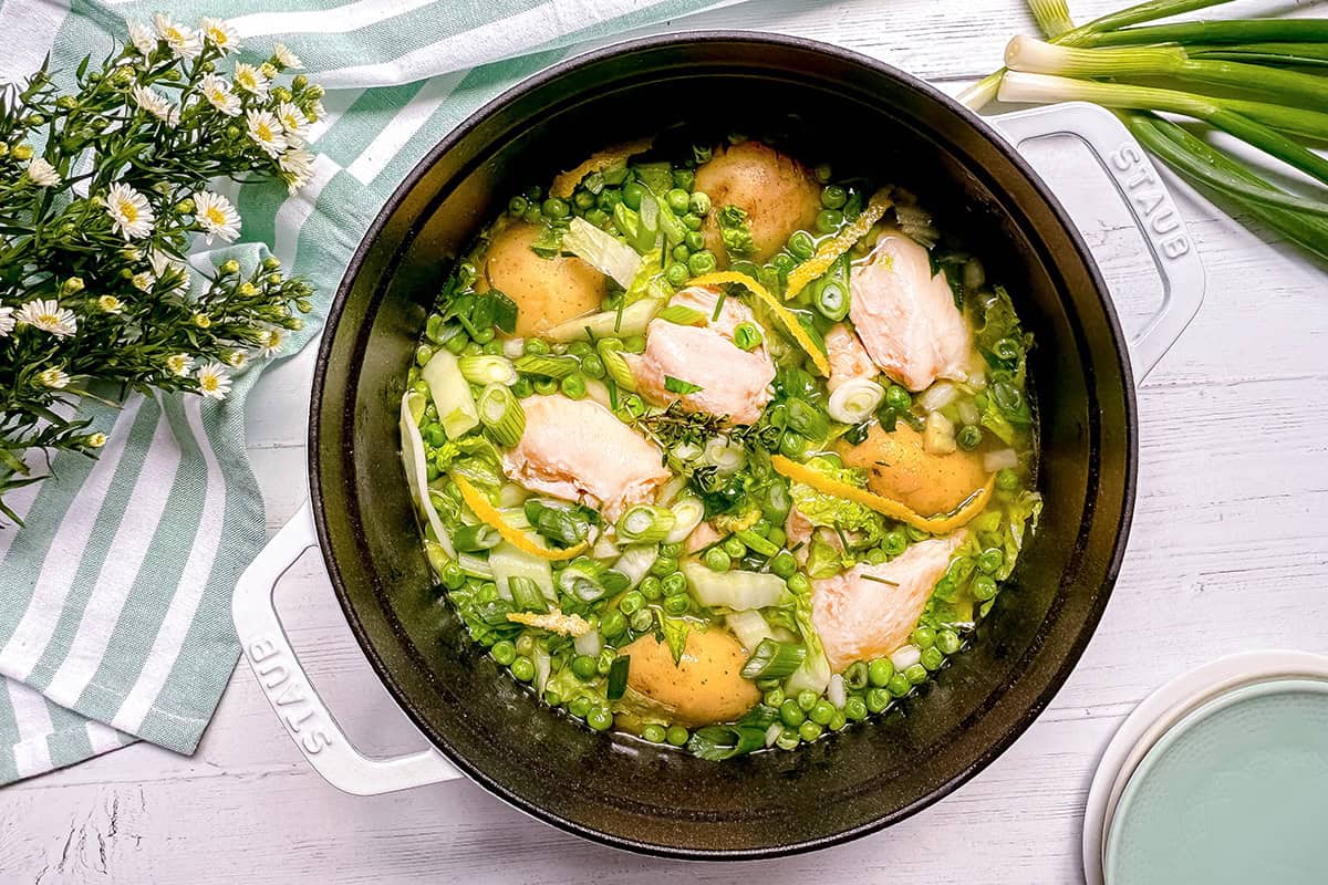 A pot of cooked Spring Chicken, potatoes, peas, and chopped green onions in broth sits surrounded by a striped cloth, flowers, and fresh green onions on a white table.
