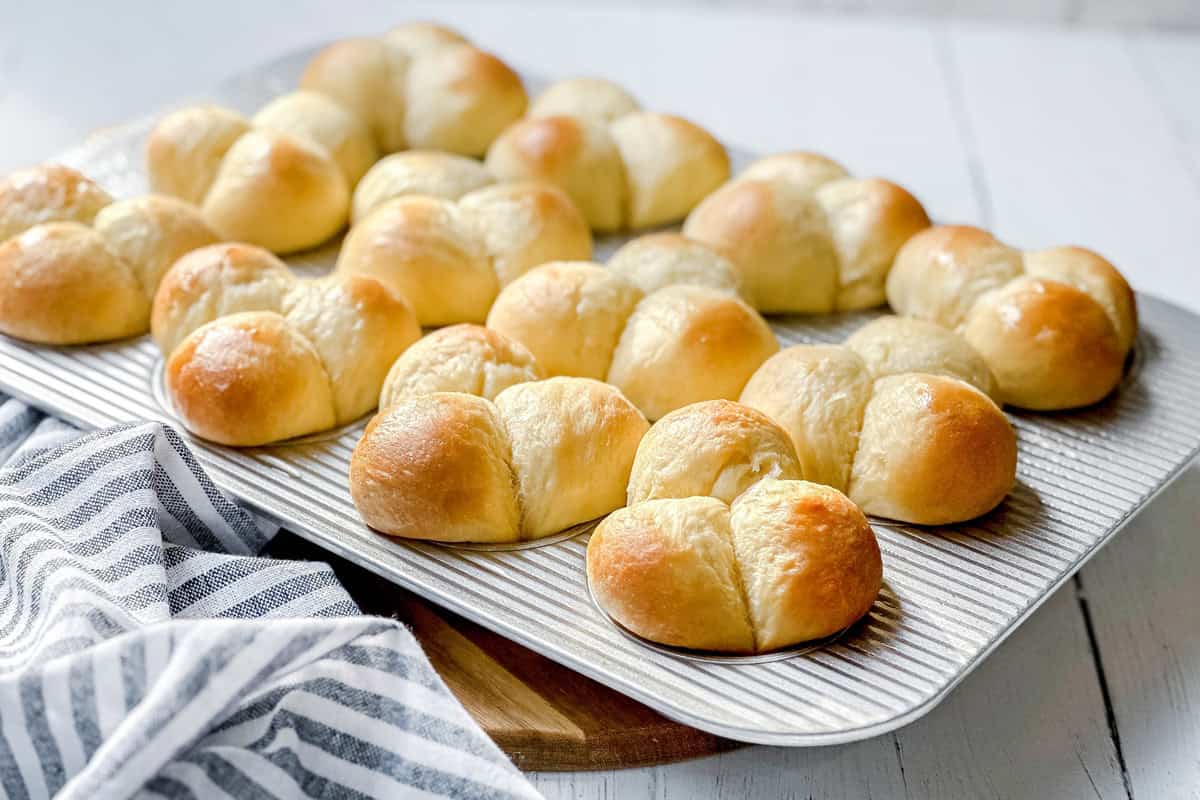 Golden dinner rolls, freshly baked, are arranged on a baking tray. The tray rests on a wooden board with a striped cloth beside it, all atop a white wooden surface. The dinner rolls look soft and have a glossy finish.