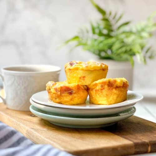 Three egg muffins on stacked plates sit on a wooden board next to a white cup. A leafy green plant and marble background are in the distance, with soft natural lighting highlighting this homemade egg bites recipe.