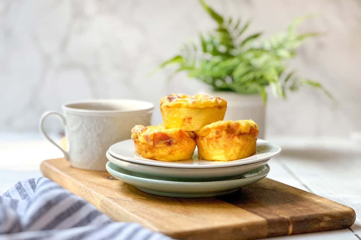 Three egg muffins on stacked plates sit on a wooden board next to a white cup. A leafy green plant and marble background are in the distance, with soft natural lighting highlighting this homemade egg bites recipe.