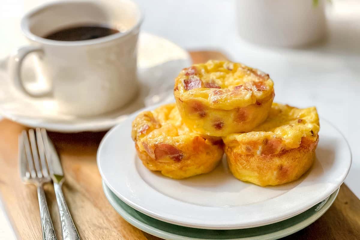 Three egg muffins with ham, reminiscent of bacon gouda egg bites, sit on a white plate next to a cup of black coffee and cutlery on a wooden serving board.