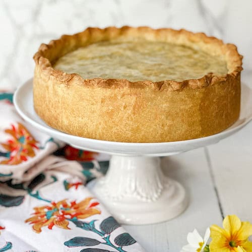 A golden-brown pie reminiscent of pizza rustica sits on a white pedestal cake stand. The pie boasts a thick crust and smooth top, while a colorful floral cloth lies beside it. Nearby, a few yellow and white flowers rest on the white wooden surface, adding to the rustic charm.