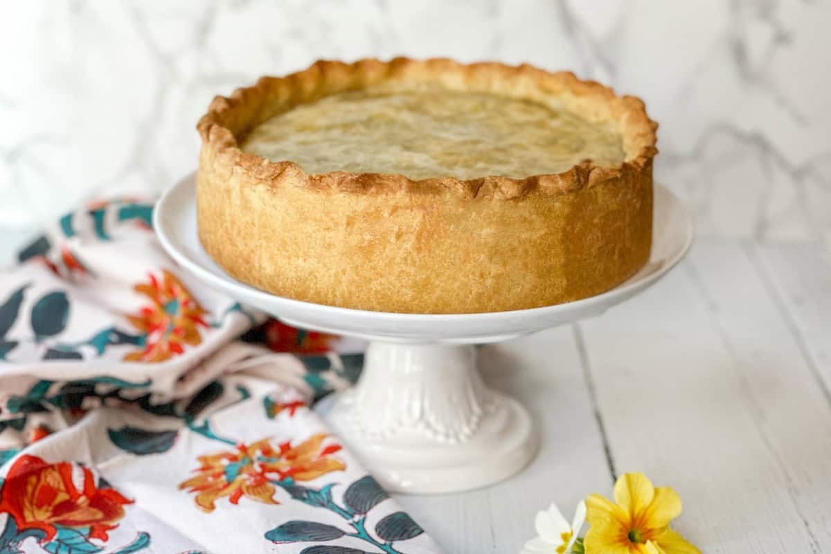 A golden-brown pie reminiscent of pizza rustica sits on a white pedestal cake stand. The pie boasts a thick crust and smooth top, while a colorful floral cloth lies beside it. Nearby, a few yellow and white flowers rest on the white wooden surface, adding to the rustic charm.