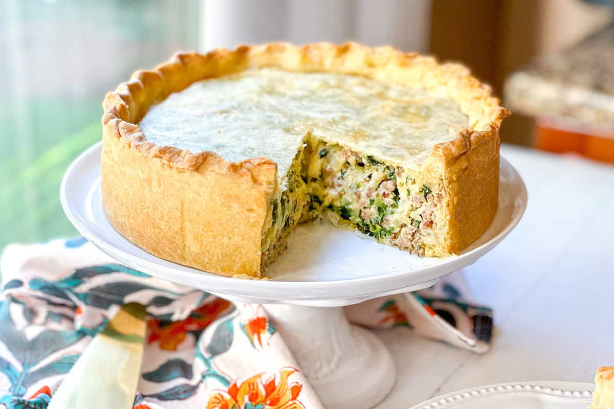 A deep-dish pizza rustica savory pie with a slice removed to reveal its hearty filling on a white cake stand.