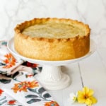 A round, golden-brown pizza rustica with a thick crust sits on a white cake stand next to a floral napkin and two small flowers.