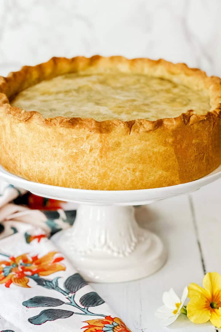 A round, golden-brown pizza rustica with a thick crust sits on a white cake stand next to a floral napkin and two small flowers.