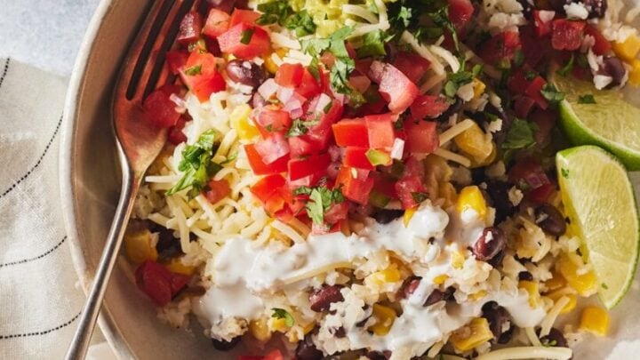 A quick April 20 minute dinner: a bowl of rice topped with black beans, corn, shredded cheese, diced tomatoes, guacamole, sour cream, chopped cilantro, and two lime wedges, served with a fork on the side.