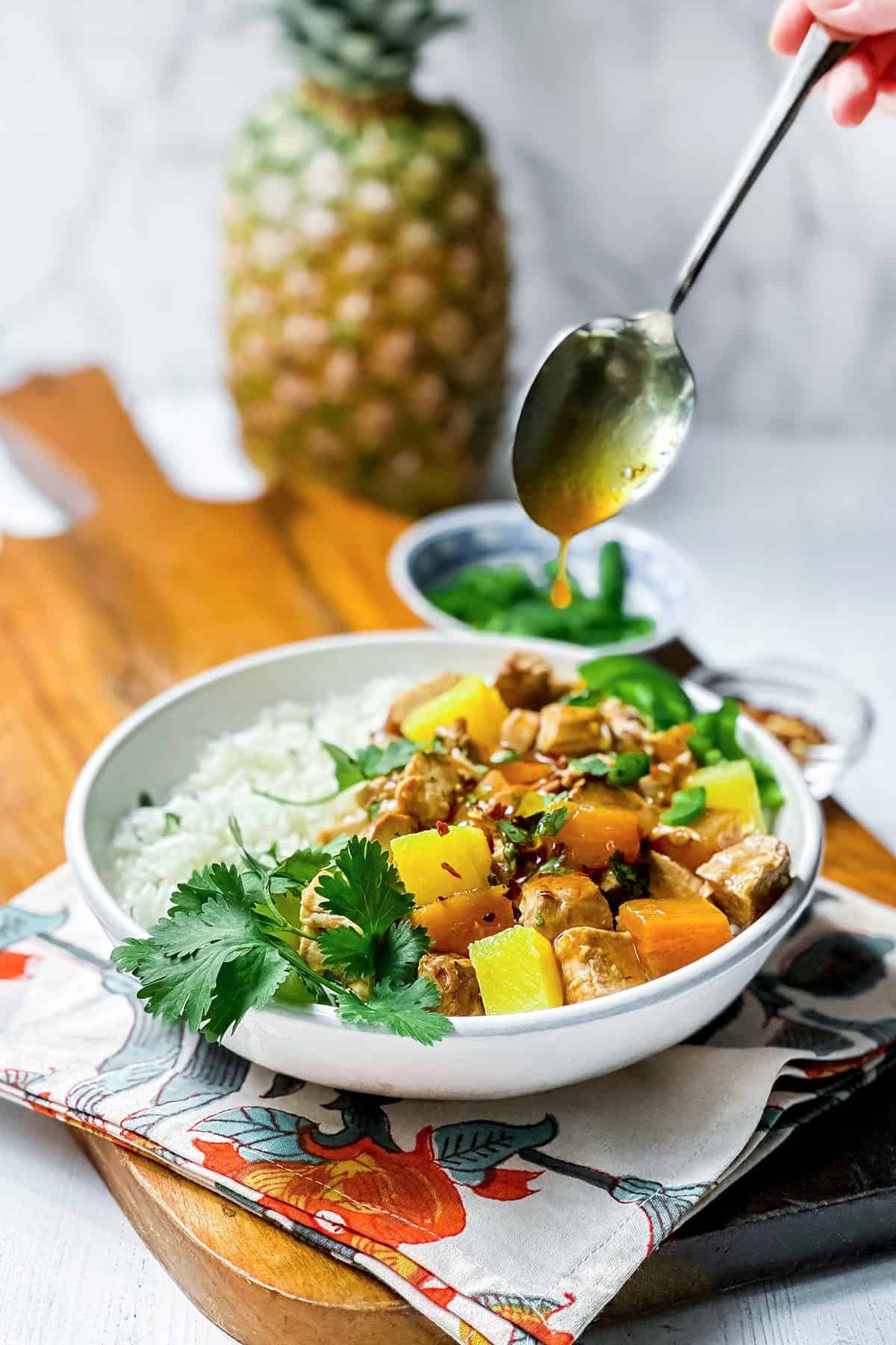 A bowl of rice and crockpot pineapple pork tenderloin garnished with cilantro, with a hand drizzling sauce over the dish; a pineapple, herbs, and savory pineapple pork are in the background.