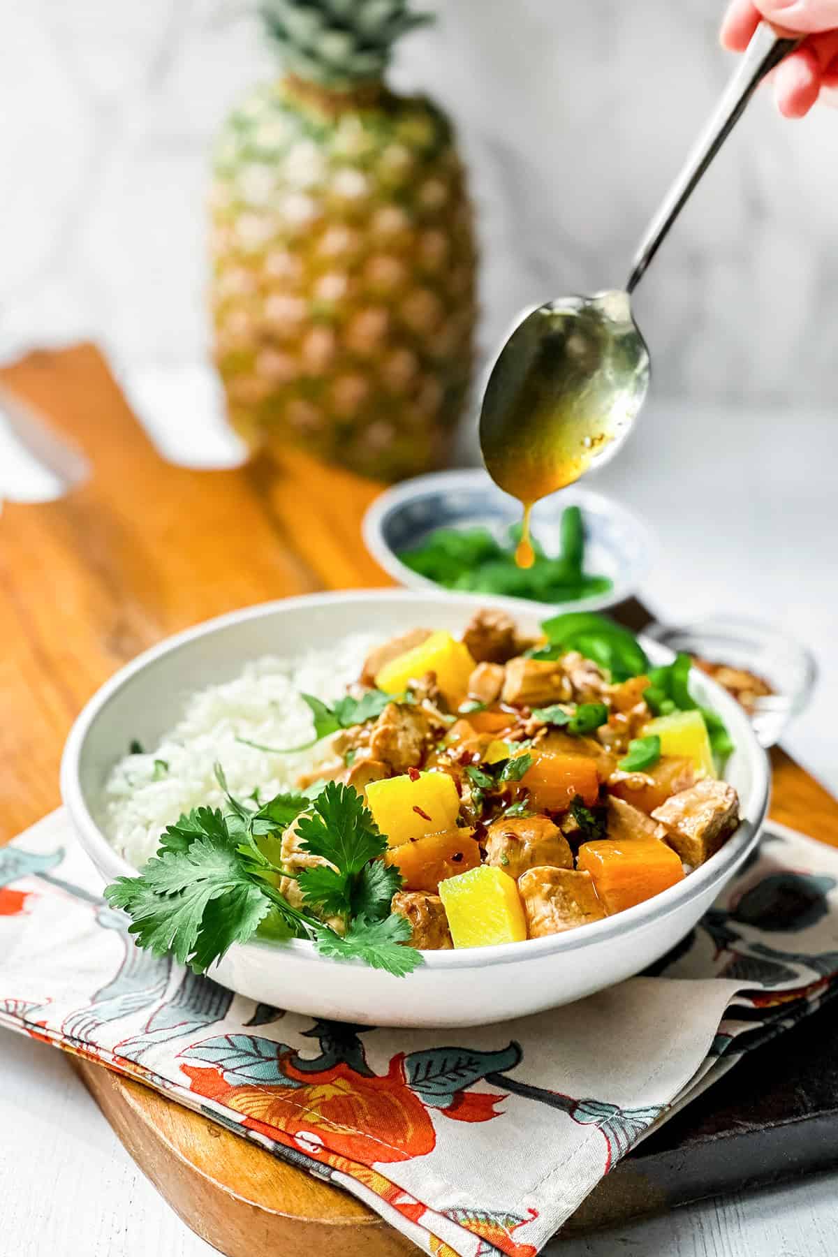 A bowl of rice and crockpot pineapple pork tenderloin garnished with cilantro, with a hand drizzling teriyaki sauce over the dish.