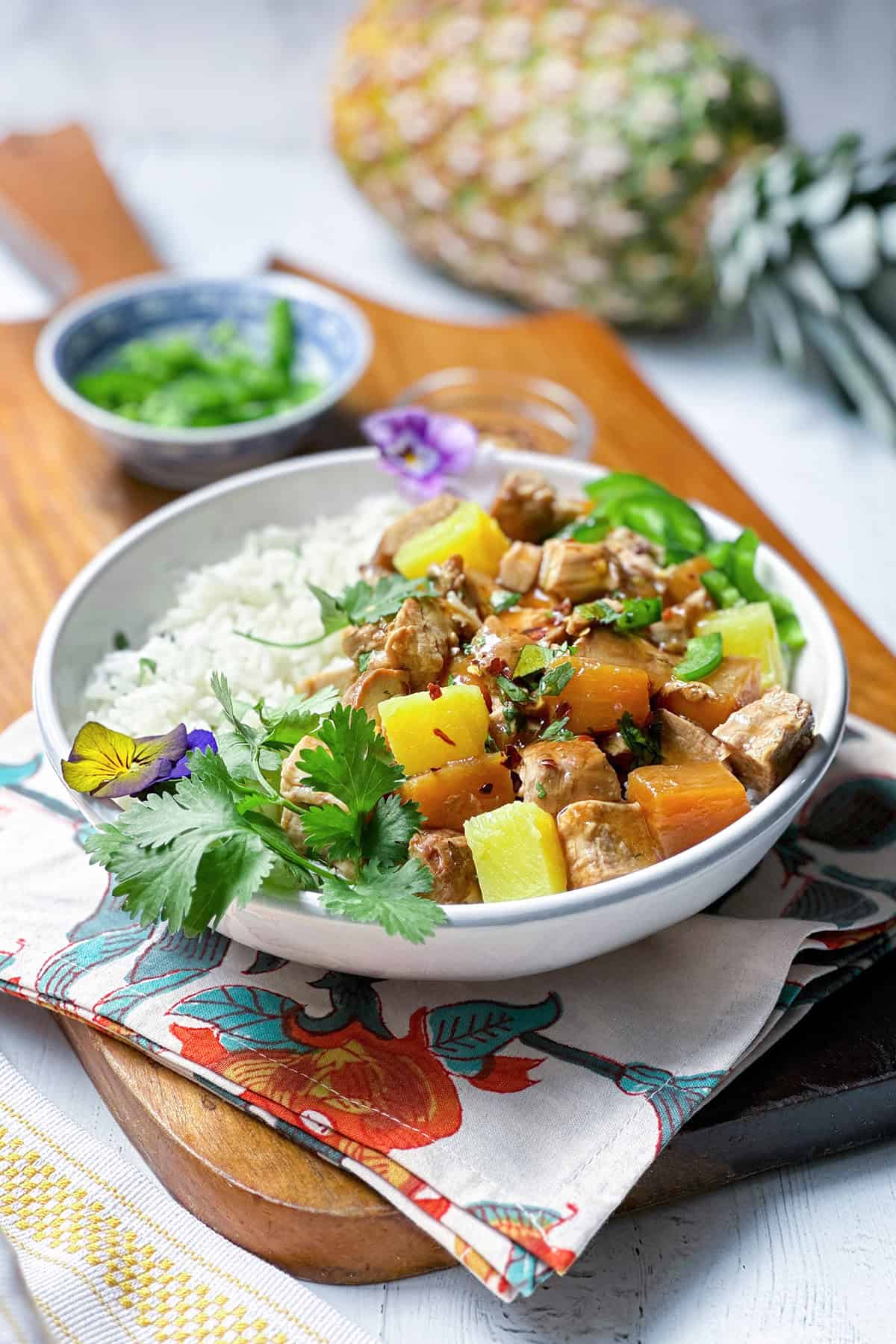 A bowl of white rice topped with crockpot Hawaiian pineapple pork tenderloin, pineapple chunks, and herbs sits on a patterned napkin, while a small bowl of chopped greens and a fresh pineapple pork dish appear in the background.