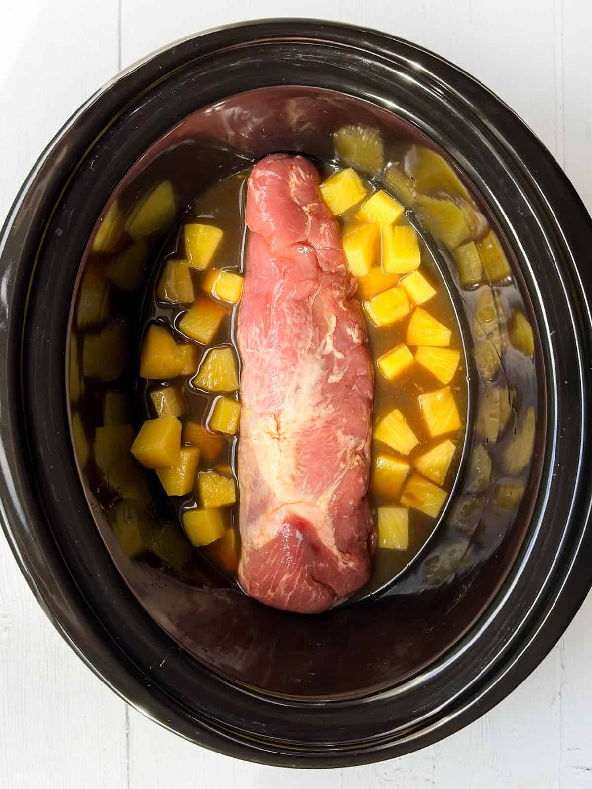 Top view of uncooked pork tenderloin in crockpot insert with teriyaki sauce and pineapples.
