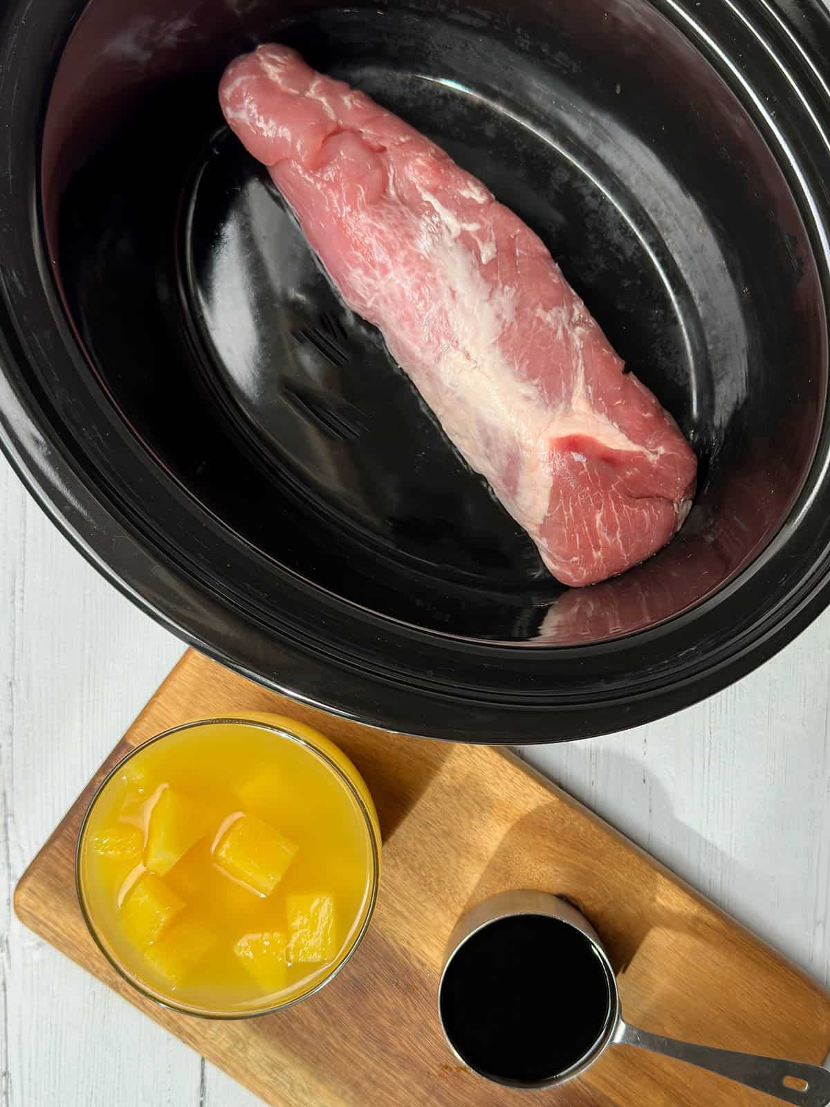 Uncooked pork tenderloin in slow cooker insert with a bowl of teriyaki sauce and pineapple chunks in their juices nearby.