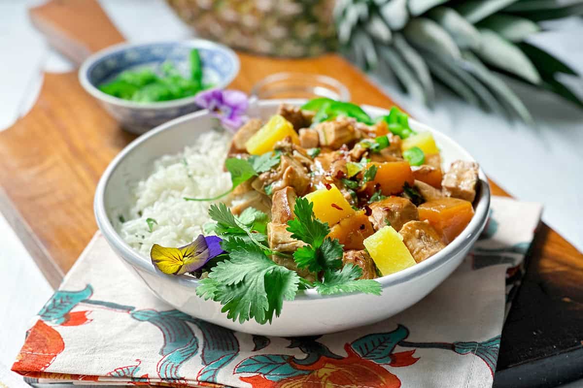 A bowl of white rice and stir-fried tofu with pineapple pork is garnished with fresh cilantro and edible flowers, sitting on a patterned napkin with a small bowl of green vegetables beside it.