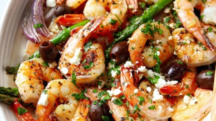 Oval dish filled with cooked shrimp, asparagus, red onions, tomatoes, black olives, and crumbled feta cheese, garnished with chopped parsley—a fresh take on Healthy Sheet Pan Dinners.