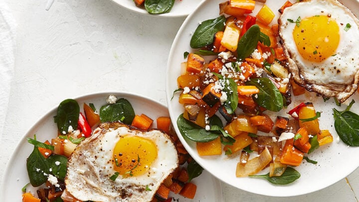 Four plates with fried eggs on top of sautéed vegetables and spinach are arranged on a white surface, showcasing a vibrant twist on Healthy Sheet Pan Dinners—each served with a fork for easy enjoyment.