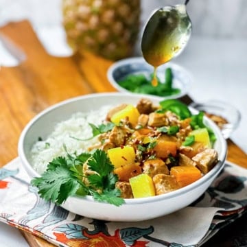 A bowl of coconut rice and pineapple pork with colorful bell peppers and pineapple is garnished with cilantro, while a savory Pineapple Pork sauce is being drizzled from a spoon above.