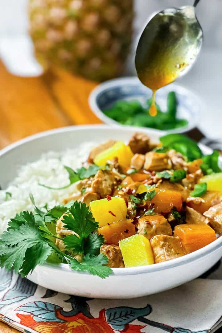A bowl of coconut rice and pineapple pork with colorful bell peppers and pineapple is garnished with cilantro, while a savory Pineapple Pork sauce is being drizzled from a spoon above.