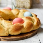 A braided round loaf of Italian Easter Bread, decorated with three colored eggs, is placed on a wooden board on a white table.