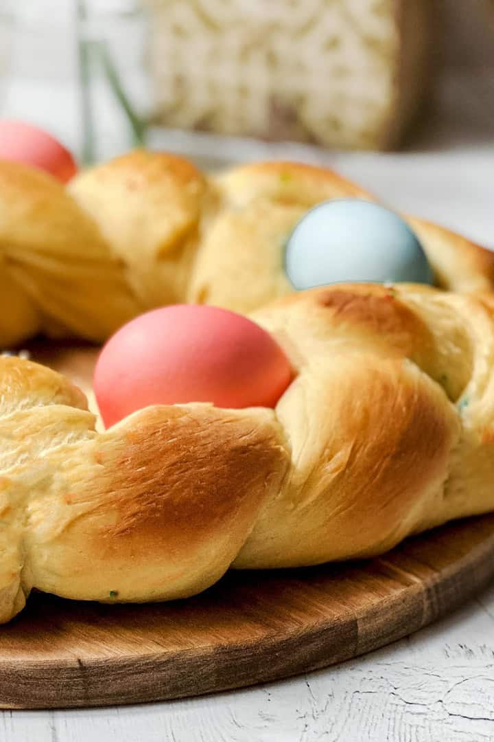 A braided round loaf of Italian Easter Bread, decorated with three colored eggs, is placed on a wooden board on a white table.
