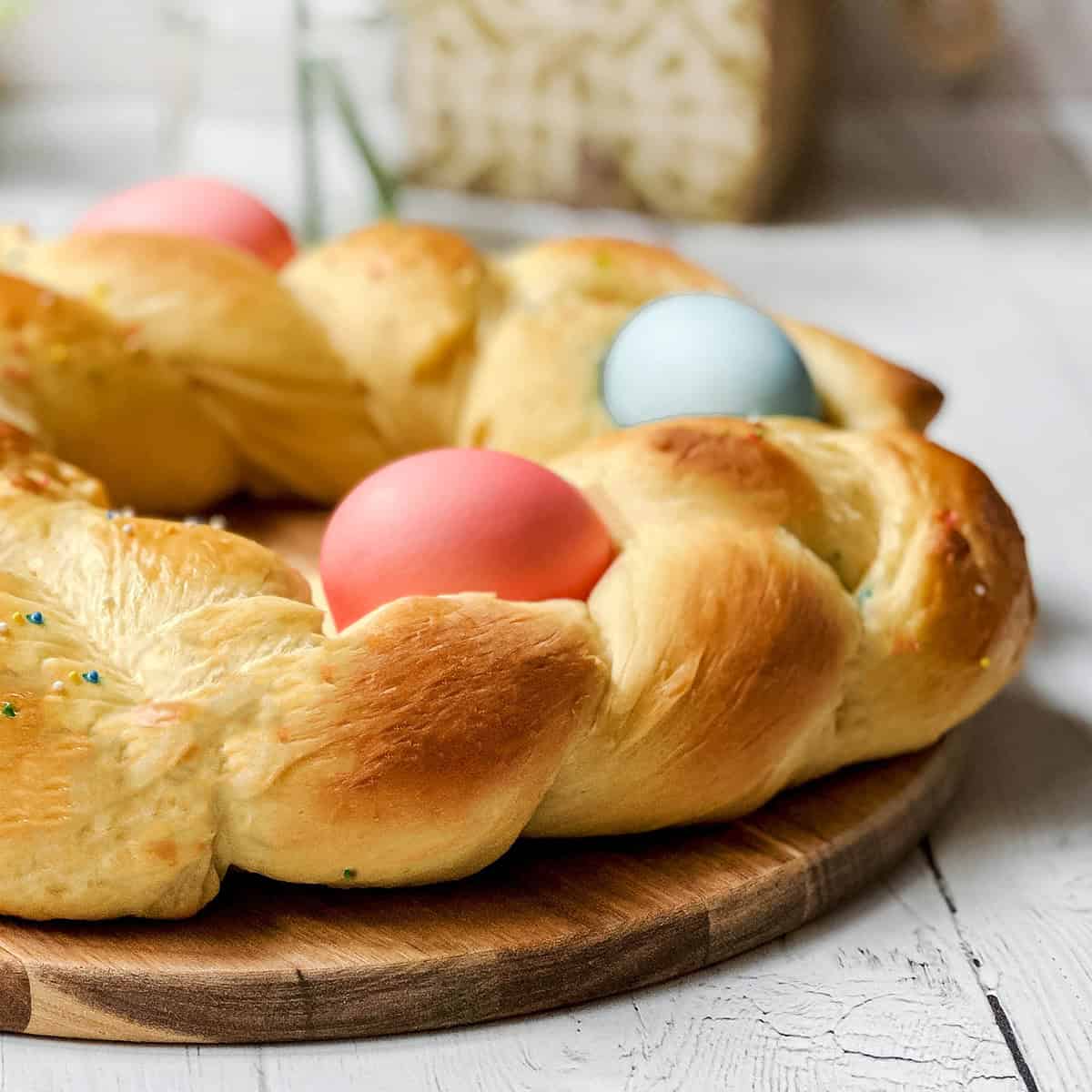 A braided round loaf of Italian Easter Bread, decorated with three colored eggs, is placed on a wooden board on a white table.