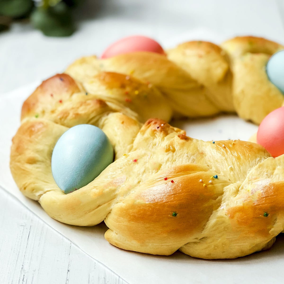 An Italian Easter Bread, braided into a ring with colorful dyed eggs and sprinkles, is displayed on a white surface.