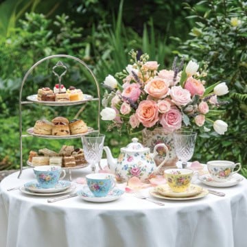 A garden table set for a Mother's Day Tea with floral china, a teapot, tiered tray of pastries, sandwiches, scones, and a bouquet of roses and tulips against a lush green background.