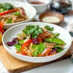 A white bowl filled with cold Curried Singapore Noodles, tossed with a colorful vegetable stir-fry of snow peas, red bell peppers, carrots, and onions, and garnished with cilantro and sesame seeds.