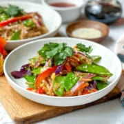 A white bowl filled with cold Curried Singapore Noodles, tossed with a colorful vegetable stir-fry of snow peas, red bell peppers, carrots, and onions, and garnished with cilantro and sesame seeds.