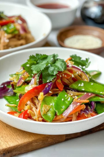 A white bowl filled with cold Curried Singapore Noodles, tossed with a colorful vegetable stir-fry of snow peas, red bell peppers, carrots, and onions, and garnished with cilantro and sesame seeds.