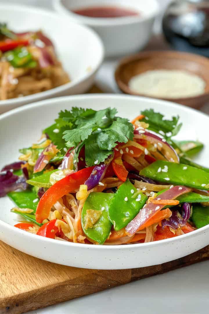 A white bowl filled with cold Curried Singapore Noodles, tossed with a colorful vegetable stir-fry of snow peas, red bell peppers, carrots, and onions, and garnished with cilantro and sesame seeds.