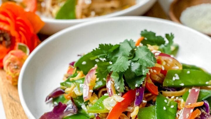 Two white plates of Vegan Curried Singapore Noodles stir-fry with snow peas, red peppers, red cabbage, onions, and cilantro, served on a wooden board with a small bowl of sauce in the background.