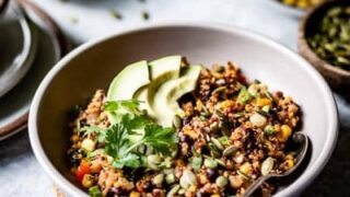 A bowl of quinoa salad topped with avocado slices and fresh parsley, perfect for April anti inflammatory dinners, with a spoon in the bowl and various ingredients in the background.