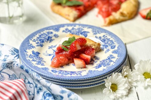 A slice of strawberry rhubarb tart galette garnished with mint sits on a blue and white patterned plate, with a whole galette and flowers in the background.