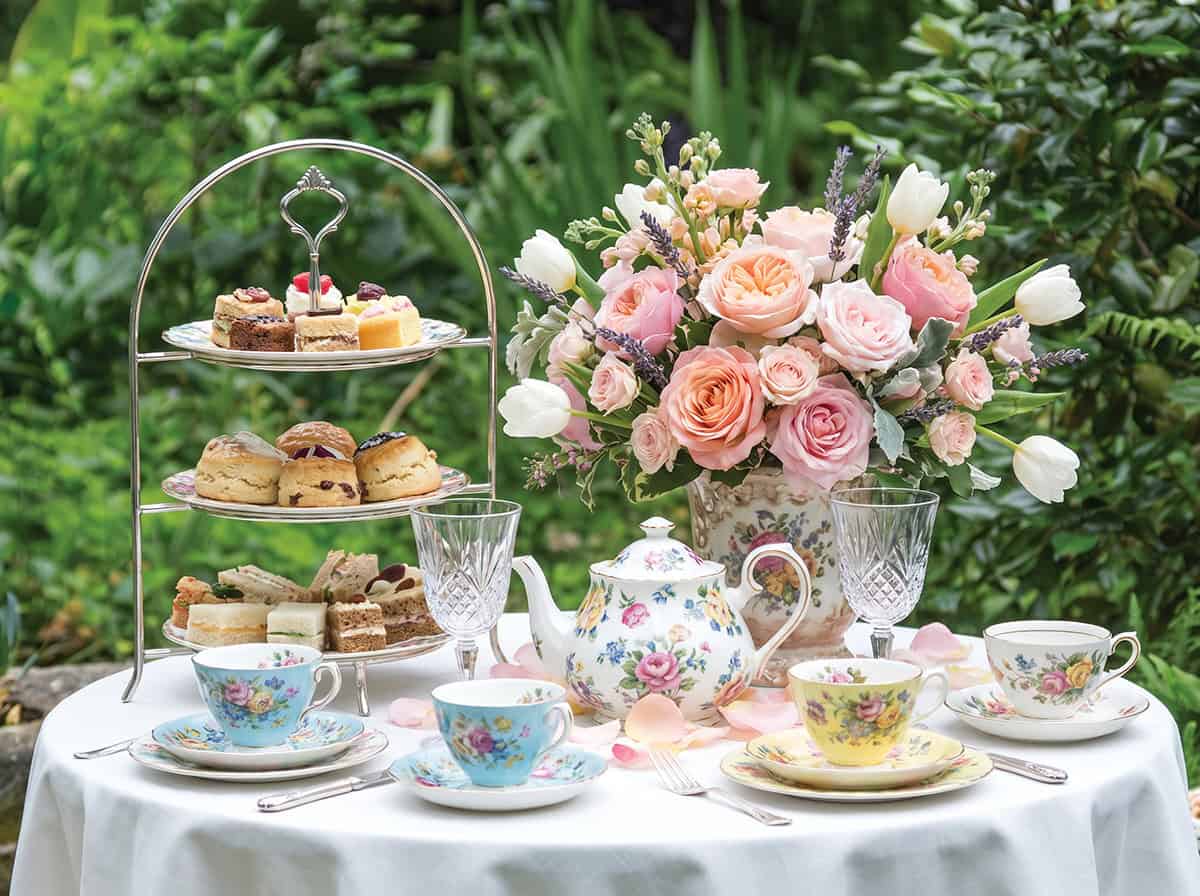 A table set for a Mother's Day Tea features floral china, a teapot, three teacups, a tiered tray of pastries, crystal glasses, and a bouquet of pink and white flowers, all arranged outdoors in a blooming garden.