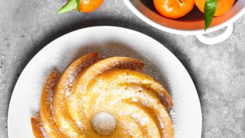 A bundt cake dusted with powdered sugar on a white plate sits next to a bowl of fresh mandarins with leaves, set on a gray surface—perfect for celebrating May Day Dinners.