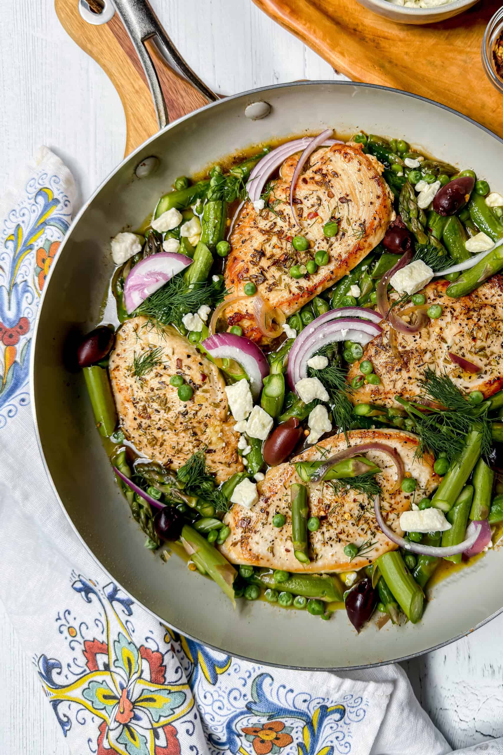A Greek Chicken skillet featuring cooked chicken breasts, asparagus, peas, red onion slices, olives, feta cheese, and fresh herbs sits on a patterned cloth.