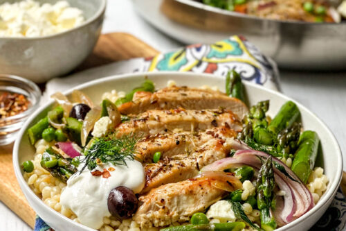 Side view of plated Greek Chicken in a white pasta bowl with fresh dill, olives, and feta with the skillet in the background