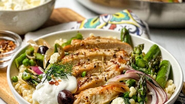 Side view of plated Greek Chicken in a white pasta bowl with fresh dill, olives, and feta with the skillet in the background