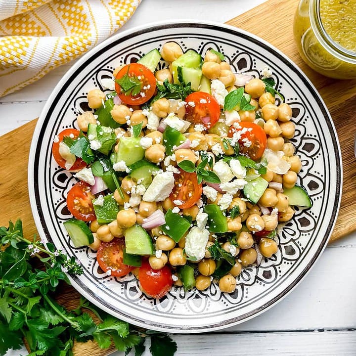 Horizontal top view of Chickpea Salad on a black and white plate with fresh herbs, cucumbers and feta cheese
