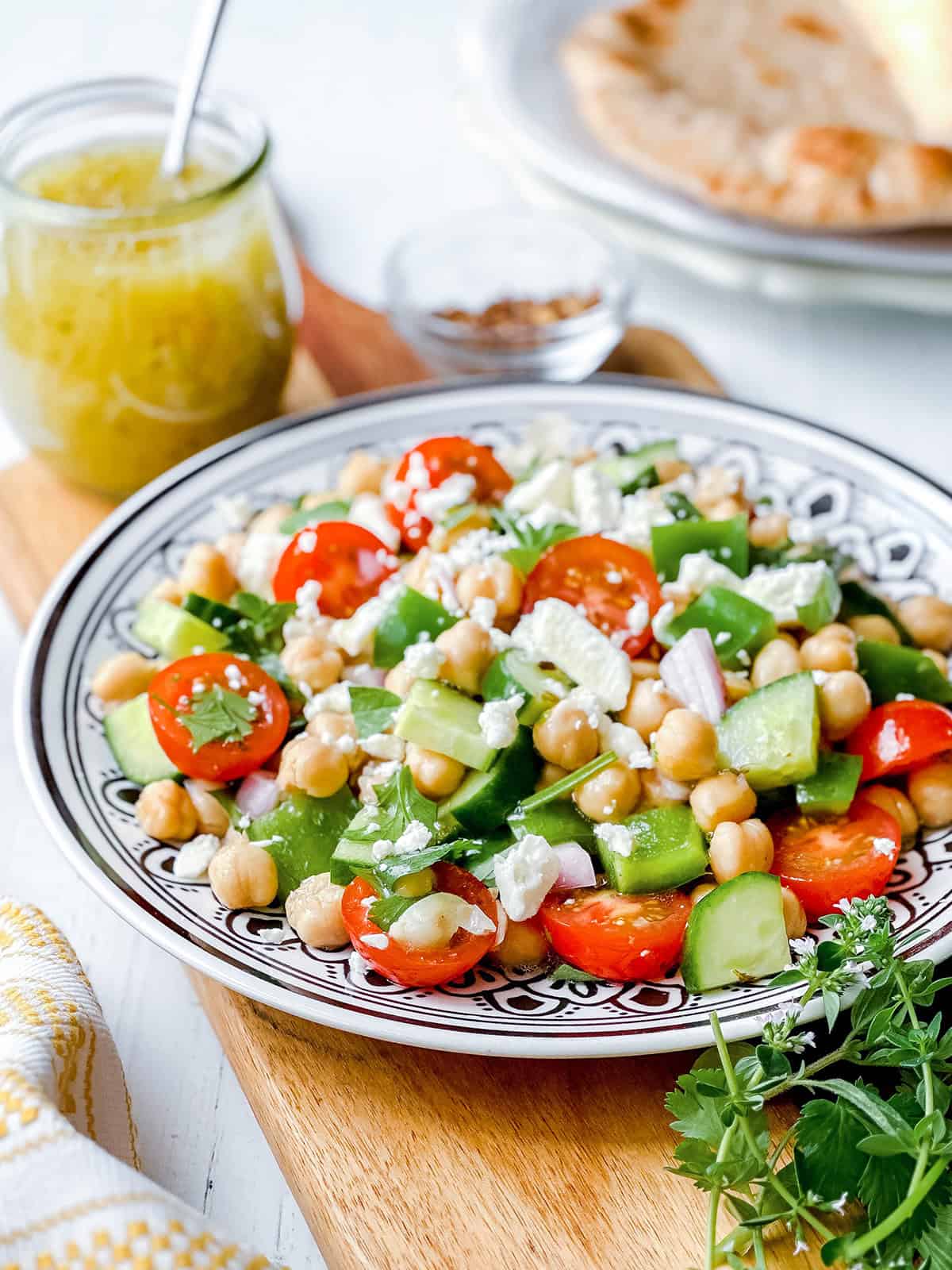 A plate of chickpea salad with cherry tomatoes, cucumber, green peppers, red onion, and feta cheese. A jar of dressing and flatbread are in the background.