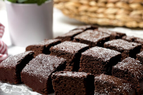 Sliced chocolate brownies, reminiscent of a rich chocolate cake, dusted with powdered sugar on parchment paper, with a potted plant and woven basket in the background.