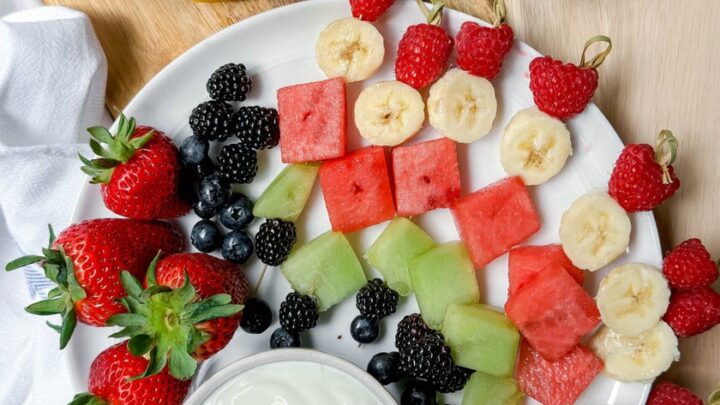 A plate with assorted fresh fruit skewers—strawberries, blackberries, blueberries—and a bowl of yogurt dip sits next to a bowl of mixed berries and cut lemons on a wooden surface.